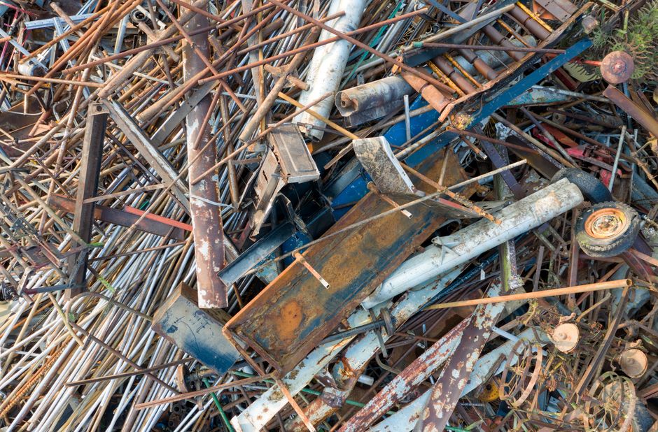 A large accumulation of discarded metal objects, including rusted pipes, sheets, rods, and frames, piled haphazardly on an outdoor surface, with some pieces showing peeling paint and corrosion. The metals vary in color from rust brown and orange to faded blue and gray, with textures ranging from smooth and bent to jagged and rough. Surrounding the pile are small patches of grass and foliage, indicating the debris is situated in an open area outside a building or yard. The scene is illuminated by natural daylight, highlighting the weathered condition of the materials. The image depicts a typical scene of scrap metal waste, which could be targeted for disposal or recycling services by companies like Rubbish Clearance Maida Vale, illustrating the type of mixed metal debris handled during alternative or private waste removal processes to prevent illegal dumping or non-compliant disposal that could lead to fines under local regulations.