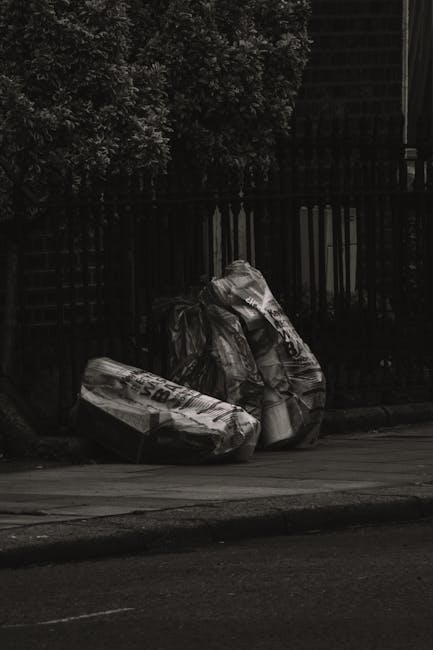 Four black plastic rubbish bags, some appearing partially torn or crumpled, are stacked against a black metal fence on a paved street. The bags are filled with waste materials and are situated near the curb, with a residential hedge and part of a building visible in the background. The overall scene is captured in low lighting, emphasizing the dark tones of the bags and surroundings, which suggests an outdoor setting typical of private rubbish disposal or collection services such as those offered by Rubbish Clearance Maida Vale, supporting alternative waste handling outside of municipal rubbish collection systems.