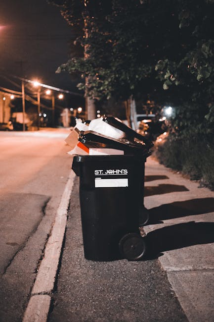 A black wheeled rubbish bin positioned on the pavement next to a curb in an urban street setting at night. The bin has a white label with the text 'ST. JOHN'S' visible on the front. The lid of the bin is open, revealing mixed waste including cardboard and plastic items. The surrounding environment features dim street lighting, with a row of illuminated street lamps in the background, tall trees with dark foliage, and a distant view of parked cars, suggesting a residential or lightly trafficked area. Shadows are cast on the pavement by the bin and nearby objects, and the scene appears quiet, with no people present. The image conveys a scene of on-site rubbish disposal possibly illustrating private waste collection or a local refuse disposal method, aligning with the theme of independent rubbish removal services provided by companies such as Rubbish Clearance Maida Vale.