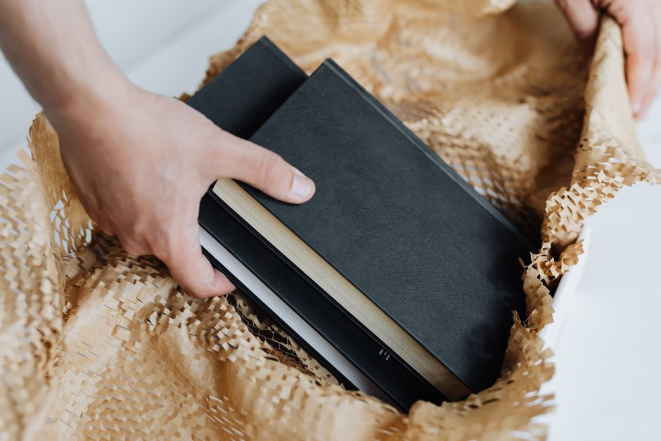 A close-up of a person's hand placing three black hardcover books into a brown paper recycling bag, which is lined with a crumpled, textured paper padding. The books are stacked with their spines facing inward, and the papers in the bag exhibit a slightly rough, fibrous surface. The scene suggests an activity related to waste disposal or recycling, possibly conducted by Rubbish Clearance Maida Vale, with the focus on separating paper materials for collection. The background surfaces are neutral and out of focus, emphasizing the action of the hand and the contents of the bag, aligning with an environmentally conscious approach to rubbish removal and private waste handling in a domestic or office setting.