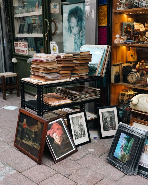 A street scene outside a shop displaying a variety of framed photographs and artworks. On a black, multi-level metal trolley, there are numerous stacks of paper, photographs, and prints, some arranged in neat piles while others are leaning against the sides of the trolley. Several framed photographs are placed on the pavement, featuring portraits and artworks, with a prominent one of a woman wearing a red headscarf in the foreground. Behind the trolley, the shop window showcases additional decorative items, vintage objects, and a poster of a woman’s face, with a small sign on the door indicating services related to telephone and communication repairs. The surrounding environment suggests an outdoor setting, possibly a flea market or antique shop front, where items are prepared for display or collection. The scene is illuminated by natural daylight, with a casual and cluttered appearance typical of on-site clearance or private collection activities, aligning with the context of independent rubbish or waste removal services provided by Rubbish Clearance Maida Vale.