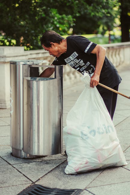 A middle-aged woman with short dark hair, dressed in a short-sleeved black t-shirt with white text and dark pants, is bending slightly forward while disposing of rubbish into a tall, cylindrical, stainless steel public trash bin with a slot opening. She is holding a large white plastic bag, filled with trash, in her left hand, and appears to be in the process of emptying it into the bin. The scene takes place outdoors on a paved area, likely in a park or public walkway, with a low stone or concrete balustrade visible in the background along with lush green trees and foliage, indicating a well-maintained urban environment. The lighting suggests daytime with natural sunlight illuminating the scene, and the overall setting conveys a focus on proper waste disposal or rubbish removal, which aligns with the services offered by Rubbish Clearance Maida Vale for private waste handling and on-site clearance activities.