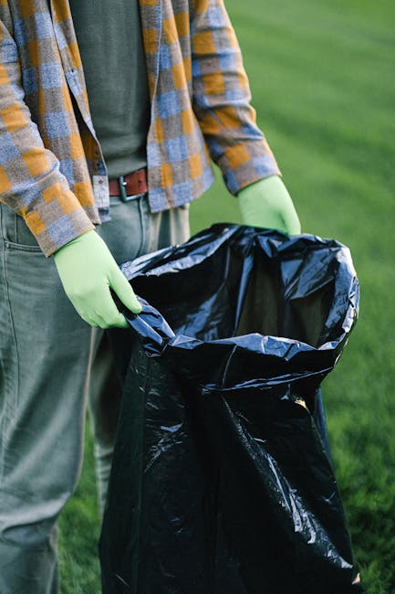 Close-up of a person wearing a yellow and gray checkered flannel shirt, gray trousers, and light green gloves, standing outdoors on a grassy area. They are holding open a large black garbage bag, which is partly folded back at the top, revealing a dark interior. The background shows a well-maintained green lawn with no visible structures. The scene suggests an outdoor waste collection or rubbish removal activity, possibly part of an on-site clearance effort facilitated by Rubbish Clearance Maida Vale, emphasizing the importance of proper waste handling and disposal outside of traditional municipal collection methods.