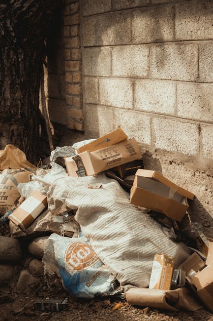 A collection of discarded cardboard boxes, some flattened and others partially intact, piled against a brick wall and a large tree trunk outdoors. The boxes are of various sizes, with some showing visible printed labels and barcodes, and they rest atop crumpled paper and a woven fabric bag. The scene is set on a patch of ground covered with small stones and dirt, with minimal natural light illuminating the area. The brick wall in the background is weathered, with uneven and aged mortar joints, suggesting an industrial or back alley environment. The presence of the rubbish in this outdoor setting reflects private or independent waste disposal, typical of a site where rubbish clearance services might be employed by Rubbish Clearance Maida Vale for efficient on-site clearance or alternative waste handling.