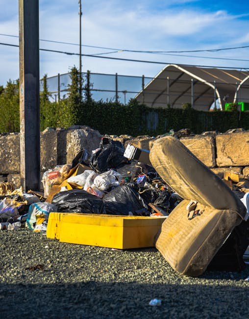 An outdoor scene showing a pile of mixed waste and rubbish dumped on a gravel surface next to a low stone wall. The waste includes black plastic bin bags, worn-out and stained upholstery from an old sofa or armchair, and a yellow plastic container. The background features a metal fence, some green foliage, and utility poles with overhead power lines against a partly cloudy sky. The image appears to demonstrate a typical example of unmanaged domestic or commercial rubbish awaiting collection or disposal, highlighting the importance of professional waste removal services such as those provided by Rubbish Clearance Maida Vale. The scene emphasizes the need for proper, sustainable disposal methods rather than illegal dumping, aligning with the context of independent and private waste handling options in the Maida Vale area. The natural lighting and outdoor environment reinforce the depiction of waste left in a public space for collection or removal, relevant to rubbish clearance or skip hire services in the vicinity.
