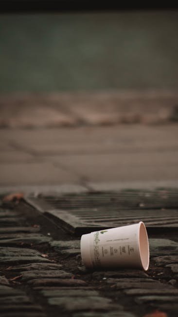 A disposable paper cup with printed text lies on a weathered brick or stone surface near a metal grate, which appears to be a drainage cover. The cup is placed on its side, partially obscuring the printed design and signs of use, such as slight dents. Surrounding the grate, the surface shows signs of dirt and possible debris, indicating an outdoor environment, possibly a pavement or driveway. In the background, a blurred, dark area suggests an outdoor setting with muted lighting, typical of an overcast day or shaded location. This scene could relate to private waste disposal or rubbish clearance services, highlighting the importance of clearing small waste items from pavements or outdoor spaces, a task often handled by professional rubbish removal companies such as Rubbish Clearance Maida Vale.