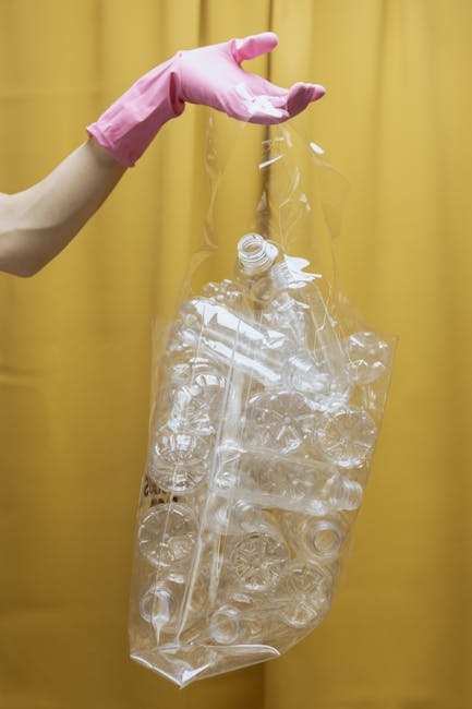 A person's hand, wearing a pink rubber glove, is holding a clear plastic bag filled with crushed and flattened transparent plastic bottles and containers. The bag appears to be partially filled, with some bottles still bearing caps and labels visible through the plastic. The background features a plain, mustard-yellow curtain or wall, providing a neutral backdrop that emphasizes the recycling activity. The scene highlights the process of sorting and preparing plastic waste for disposal or recycling, which is relevant to independent rubbish removal services offered by companies like Rubbish Clearance Maida Vale. The focus on plastic waste and the use of protective gloves suggest an emphasis on safe and environmentally conscious rubbish handling practices, aligning with professional waste management activities that promote responsible disposal and recycling efforts in the context of private waste clearance.