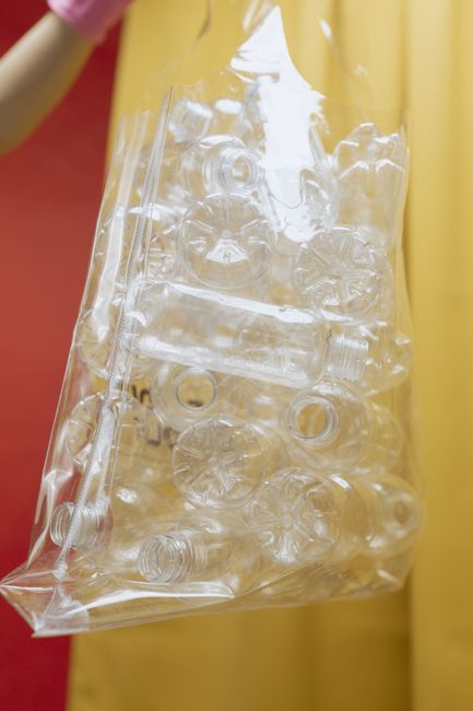 A person's hand, wearing a pink rubber glove, is holding a clear plastic bag filled with crushed and flattened transparent plastic bottles and containers. The bag appears to be partially filled, with some bottles still bearing caps and labels visible through the plastic. The background features a plain, mustard-yellow curtain or wall, providing a neutral backdrop that emphasizes the recycling activity. The scene highlights the process of sorting and preparing plastic waste for disposal or recycling, which is relevant to independent rubbish removal services offered by companies like Rubbish Clearance Maida Vale. The focus on plastic waste and the use of protective gloves suggest an emphasis on safe and environmentally conscious rubbish handling practices, aligning with professional waste management activities that promote responsible disposal and recycling efforts in the context of private waste clearance.