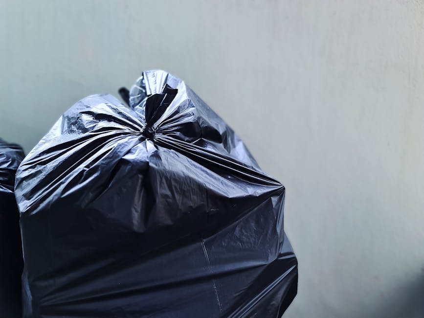 Close-up image of several black plastic rubbish bags, tightly knotted at the top, placed against a plain, light-colored wall. The bags appear to be made of shiny, slightly crinkled plastic material, with visible creases and folds. They are stacked near each other, with one bag prominently in the foreground, occupying the left half of the image. The environment suggests an indoor or enclosed space, possibly a storage area or outdoor corner adjacent to a property. The scene reflects typical waste collection preparation, consistent with private rubbish removal or alternative waste handling services offered by companies like Rubbish Clearance Maida Vale, especially when moving away from council provisions. The overall composition emphasizes the act of bagging and securing rubbish for disposal or removal, aligning with professional rubbish clearance practices in Maida Vale.