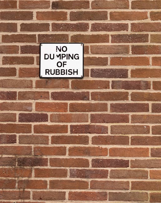 A rectangular white metal sign mounted on a red-brown brick wall with a slightly rough texture. The sign features black text that reads 'NO DUMPING OF RUBBISH,' with the words arranged in three lines, the word 'DUMPING' partially obscured or missing its first letter. The brick wall has a traditional running bond pattern, with evenly spaced horizontal mortar joints and some variation in the shade of red and brown bricks, giving a natural, weathered appearance. The wall extends vertically and horizontally beyond the edges of the image, with direct natural lighting casting a soft shadow beneath the sign, indicating daylight. The scene suggests a location in an urban or residential area where waste management and rubbish removal regulations are relevant, and the sign indicates an area where illegal dumping is discouraged. The overall setting emphasizes the importance of controlled waste disposal, aligning with services offered by companies like Rubbish Clearance Maida Vale, which provide alternative waste handling solutions outside of local council rubbish rules.