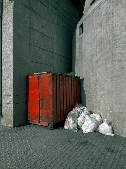 A rusty red and brown metal skip is positioned against the corner of a building, with its front facing the viewer. The skip's surface shows signs of wear, including patches of rust and chipped paint, indicating frequent use for waste collection. Surrounding the base of the skip are several plastic bags filled with mixed rubbish, including papers and recyclable materials, some of which are loosely tied or partially open. The scene is set outdoors, with the skip placed on a dark, textured pavement composed of interlocking bricks. Behind the skip, the wall on the left is clad in dark green, smooth panels, while the adjacent wall on the right is light gray with a rough, concrete-like texture. A narrow aperture or window is visible on the green wall above the skip. The lighting is natural, suggesting daytime, and there are no other objects visible in the scene, emphasizing the setting as a typical on-site rubbish clearance area, possibly used for private or independent waste disposal services such as those offered by Rubbish Clearance Maida Vale, to facilitate access in tight or restricted urban environments.