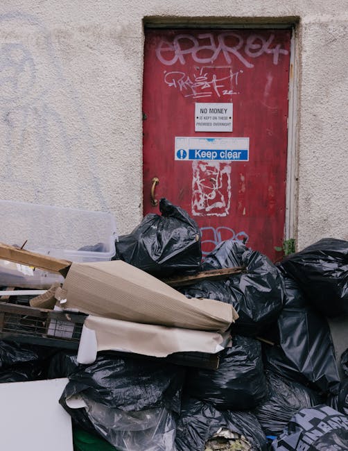 A scene of excessive rubbish accumulation in front of a red metal door set into a rough-textured, off-white concrete wall. The rubbish consists of black plastic garbage bags, some torn open, revealing discarded waste inside. Additional waste includes flattened cardboard boxes, a piece of white foam, and a partially visible metal trolley frame. The rubbish is piled directly on the pavement, which appears to be a narrow alley or urban pedestrian walkway. The door has graffiti tags in white and pink spray paint, along with a small white sign indicating 'No Money' and a blue and white sign that reads 'Keep clear.' The lighting is diffuse, suggesting an overcast day. This scene exemplifies the need for private waste collection or rubbish clearance services like those offered by Rubbish Clearance Maida Vale, especially in situations where waste cannot be easily accessed by standard council collection methods, highlighting the importance of professional rubbish removal for tight access areas.