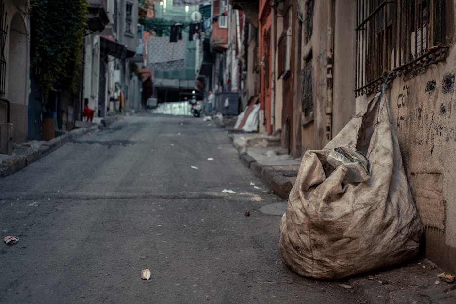 A rusty red and brown metal skip is positioned against the corner of a building, with its front facing the viewer. The skip's surface shows signs of wear, including patches of rust and chipped paint, indicating frequent use for waste collection. Surrounding the base of the skip are several plastic bags filled with mixed rubbish, including papers and recyclable materials, some of which are loosely tied or partially open. The scene is set outdoors, with the skip placed on a dark, textured pavement composed of interlocking bricks. Behind the skip, the wall on the left is clad in dark green, smooth panels, while the adjacent wall on the right is light gray with a rough, concrete-like texture. A narrow aperture or window is visible on the green wall above the skip. The lighting is natural, suggesting daytime, and there are no other objects visible in the scene, emphasizing the setting as a typical on-site rubbish clearance area, possibly used for private or independent waste disposal services such as those offered by Rubbish Clearance Maida Vale, to facilitate access in tight or restricted urban environments.